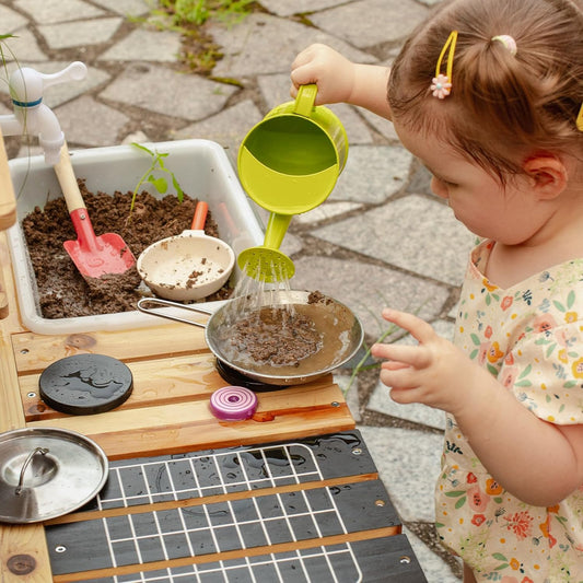 Niña jugando con cocinita de jardín experimentando juego simbólico con agua y tierra.